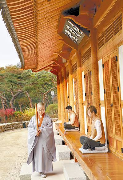 A monk in grey robes walking past a wooden temple veranda where two visitors sit on cushions